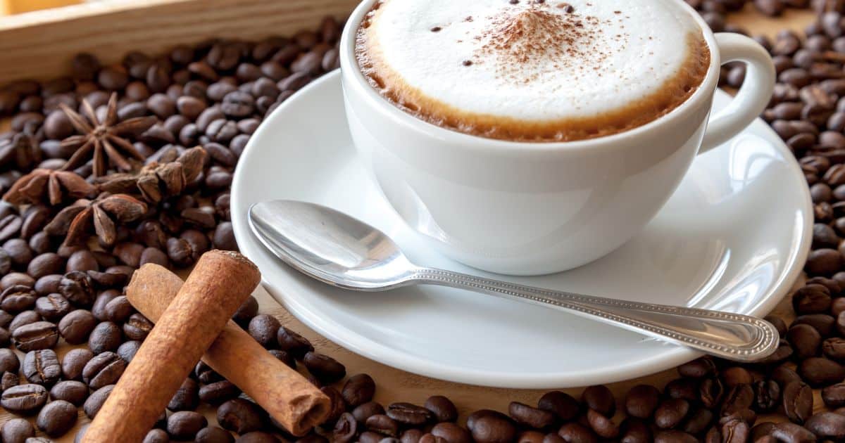 Cinnamon Cappuccino on a bed of coffee beans with two cinnamon sticks in the forefront 